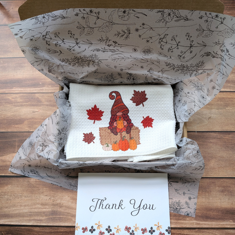 A white waffle kitchen towel with a girl fall gnome sitting in a box. Below the box is a white thank-you card.