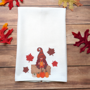 A girl gnome in a brown and orange striped hat sitting on a hay bale waffle towel. She is holding an orange pumpkin.