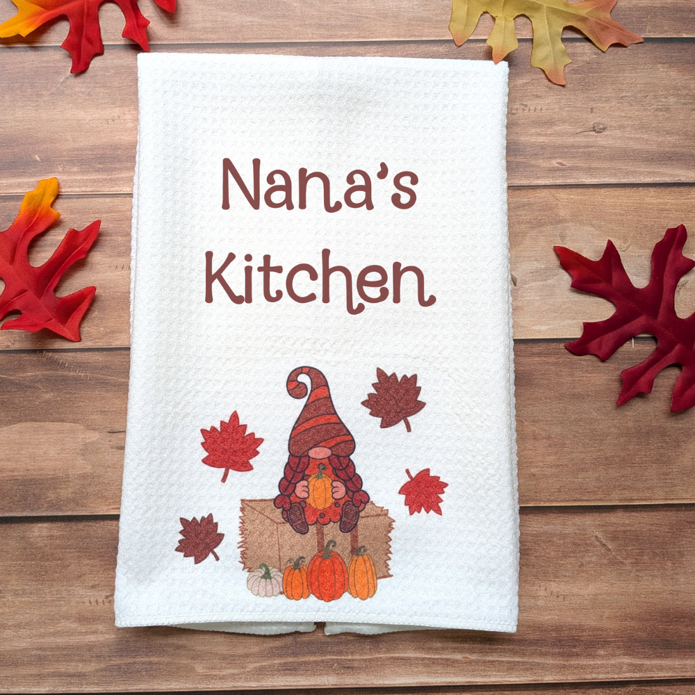 A white waffle kitchen towel with a fall girl gnome holding a pumpkin while sitting on a hay bale. The text above the gnome reads "Nana's Kitchen."