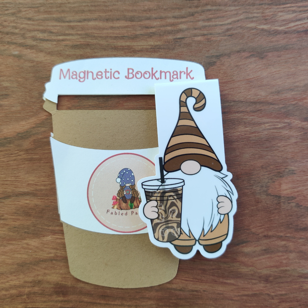 A iced coffee gnome magnentic bookmark next to coffee cup backing card. The gnome is wearing a brown striped hat and is holding an iced coffee.
