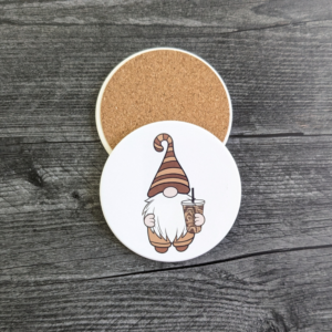 A white coaster with a gnome wearing a brown striped hat and holding an iced coffee. A back view of the coaster showing its protective cork backing.