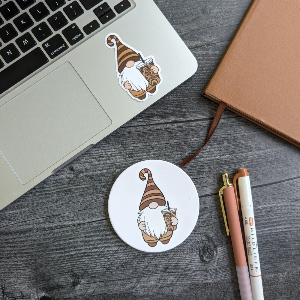 A desk with a laptop featuring an iced coffee gnome sticker. Below the laptop is the iced coffee gnome coaster.