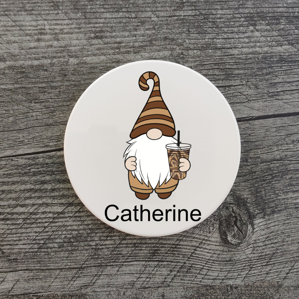 A white coaster feating a gnome in a brown striped hat and holding an iced coffee. The coaster is personalized with the name "Catherine."