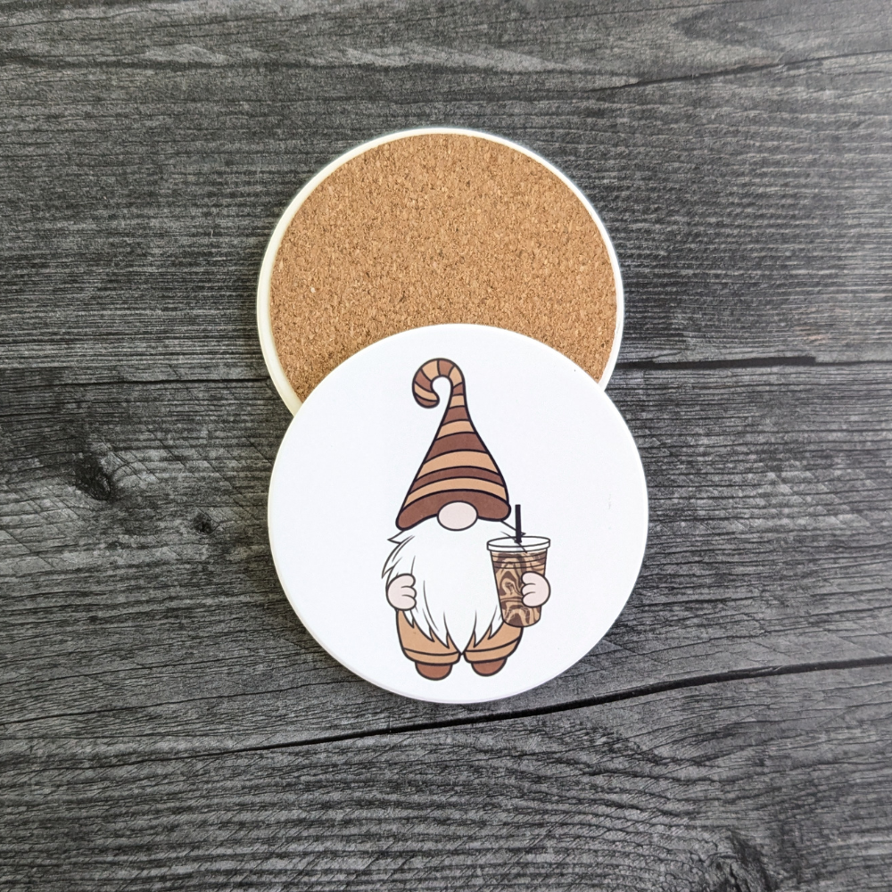 A white coaster with a gnome wearing a brown striped hat and holding an iced coffee. A back view of the coaster showing its protective cork backing.