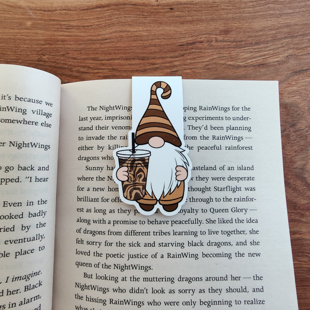 A gnome wearing a brown striped hat is holding an iced coffee magnetic bookmark. The bookmark is attached to a page in an open book.