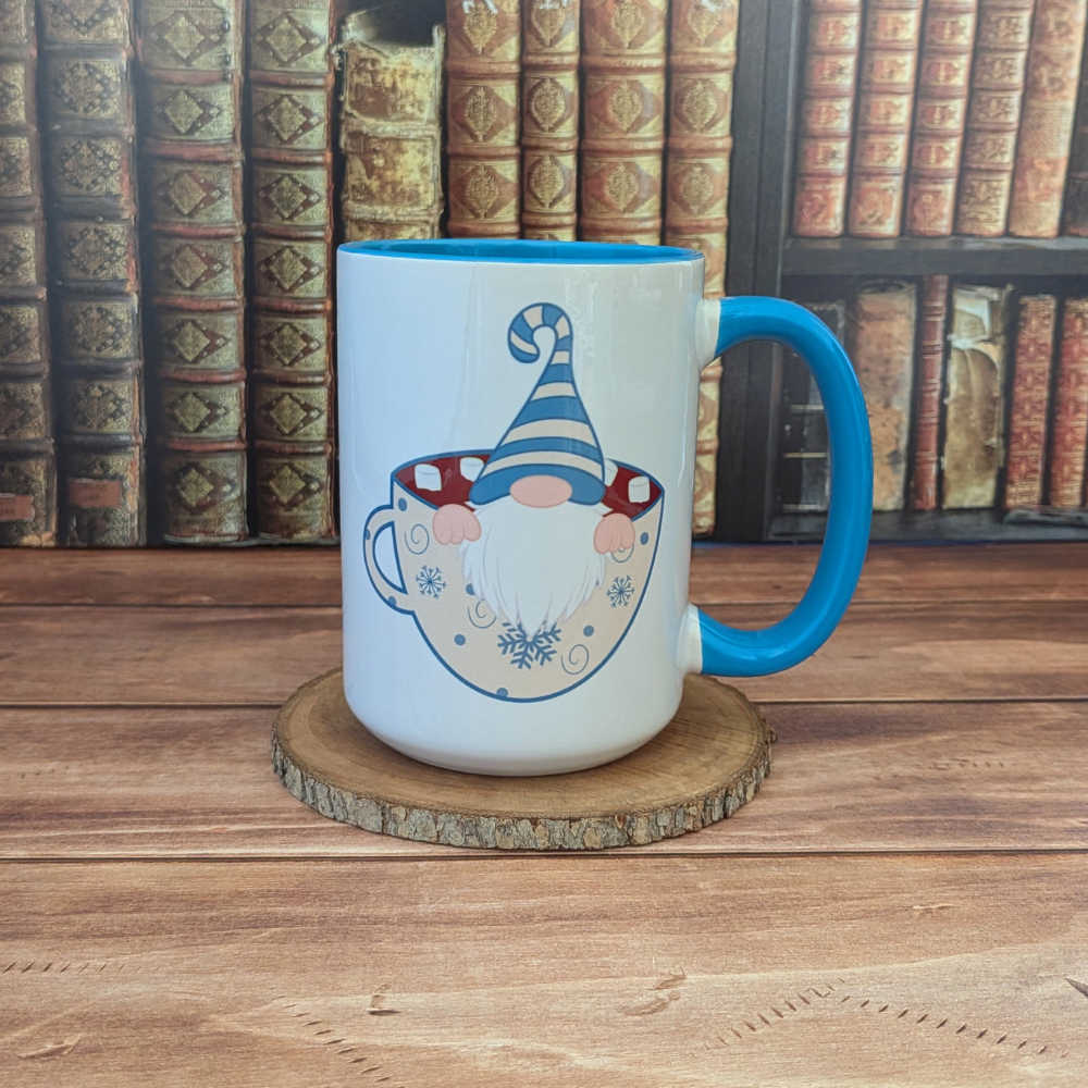 A white ceramic mug with a blue handle featuring a gnome sitting in a cup of hot chocolate. The gnome is wearing a blue-and-cream striped hat.