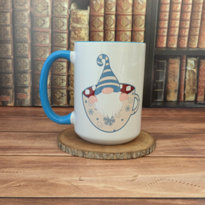 A white coffee mug with a blue handle. A gnome in a blue striped hat sitting in a cup is in the front.
