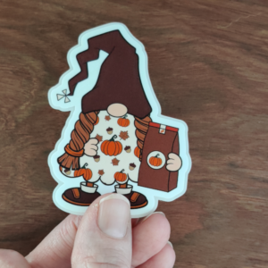 A hand holding a girl gnome sticker wearing a pumpkin fall dress while holding a pumpkin coffee bag. The sticker has a clear laminate.