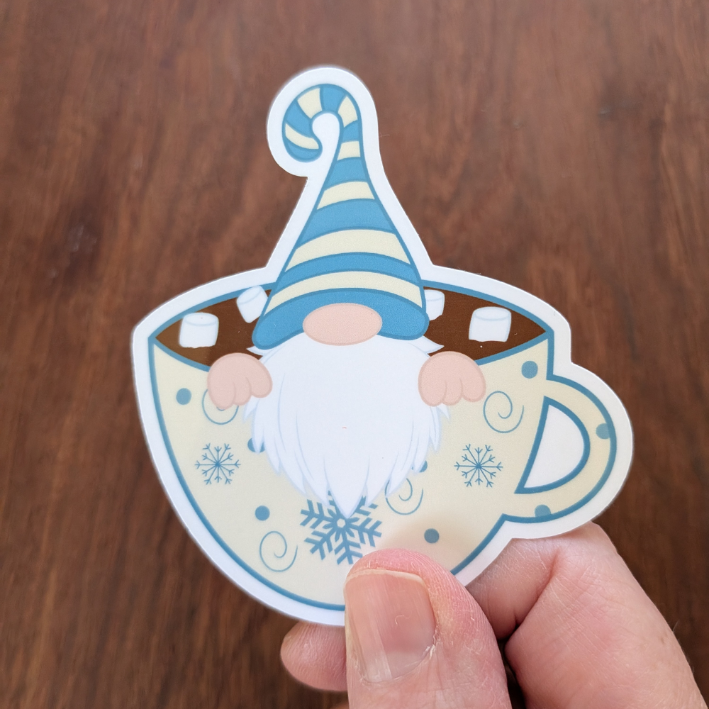 A hand holding a winter gnome sticker. The gnome is wearing a blue striped hat and is sitting in a cup of hot chocolate. The sticker has a clear laminate.