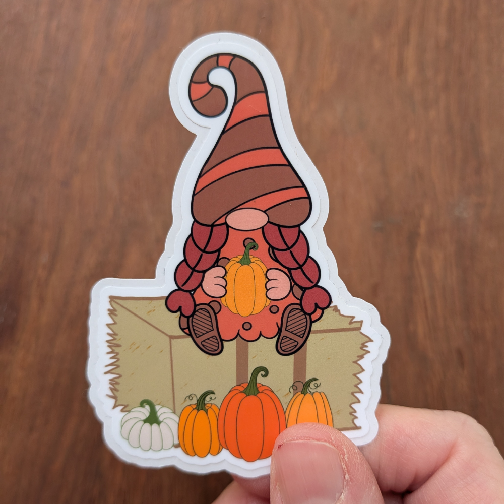 A hand holding a pumpkin patch girl gnome sticker. The sticker has a clear laminate.