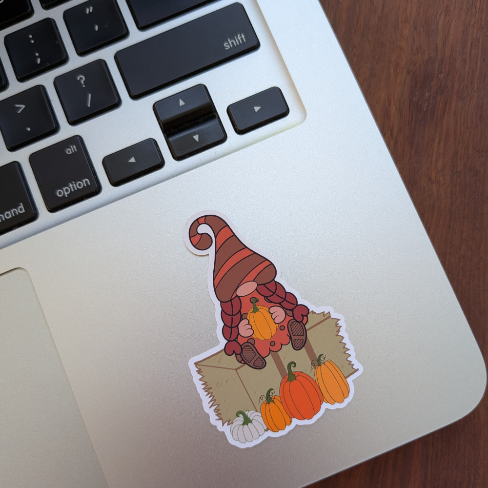 A fall girl gnome sitting on a hay bale with a pumpkin sticker. The sticker is on a silver laptop.