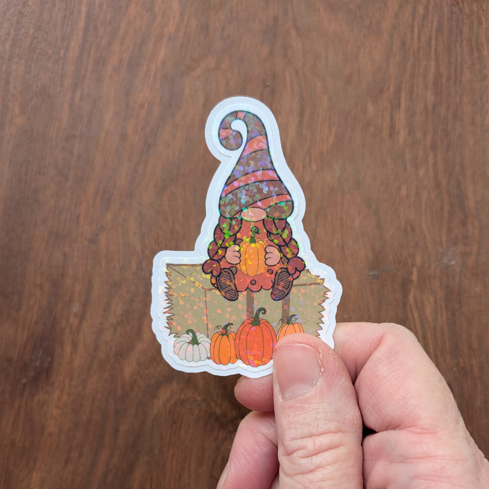 A hand holding a pumpkin patch girl gnome sticker. The sticker has a sparkle laminate.