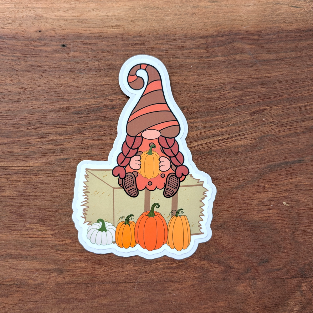A die-cut sticker featuring a gnome with a brown-and-orange striped hat, sitting on a hay bale while holding a small orange pumpkin.