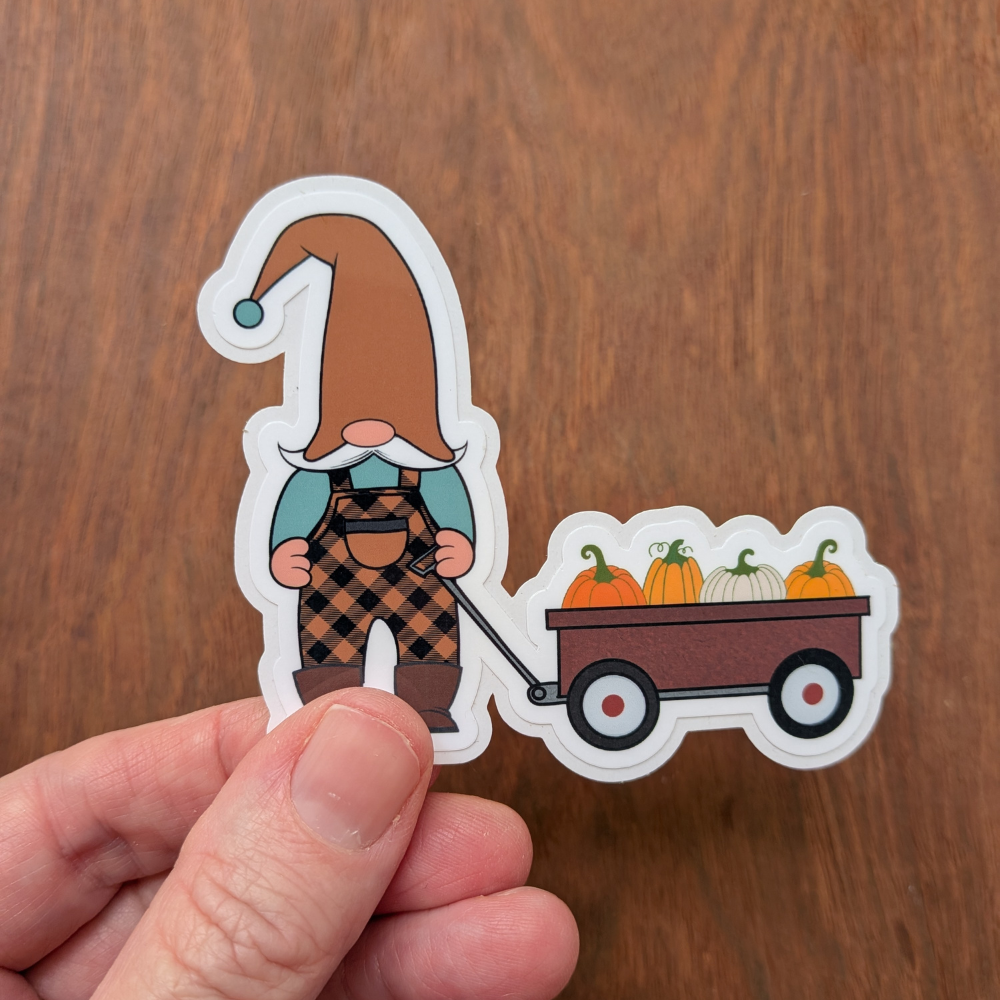 A hand holding a gnome sticker wearing orange and black overalls. The sticker has a clear laminate.