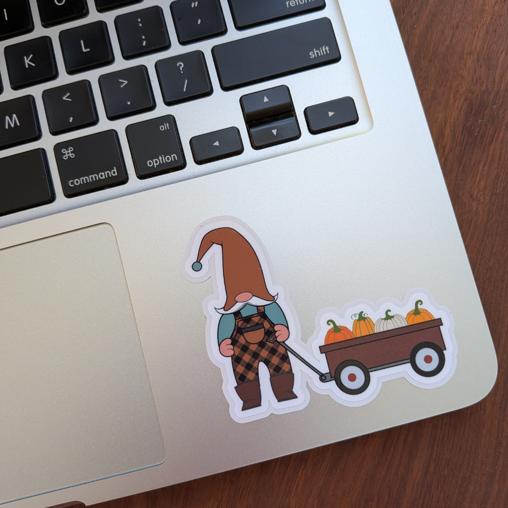 A fall gnome pulling a wagon full of pumpkins sticker. The sticker is on a silver laptop.
