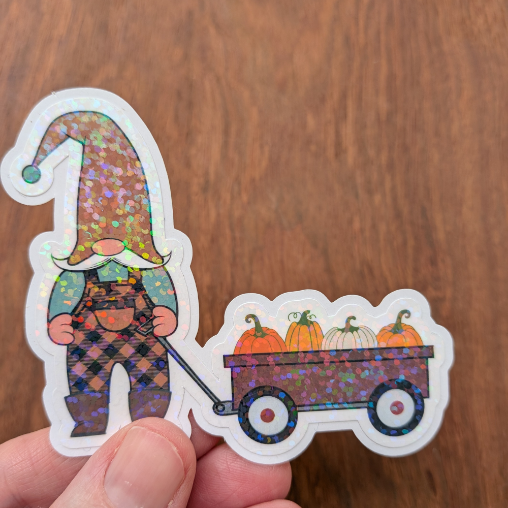 A hand holding a gnome sticker wearing orange and black overalls. The sticker has a sparkle laminate.