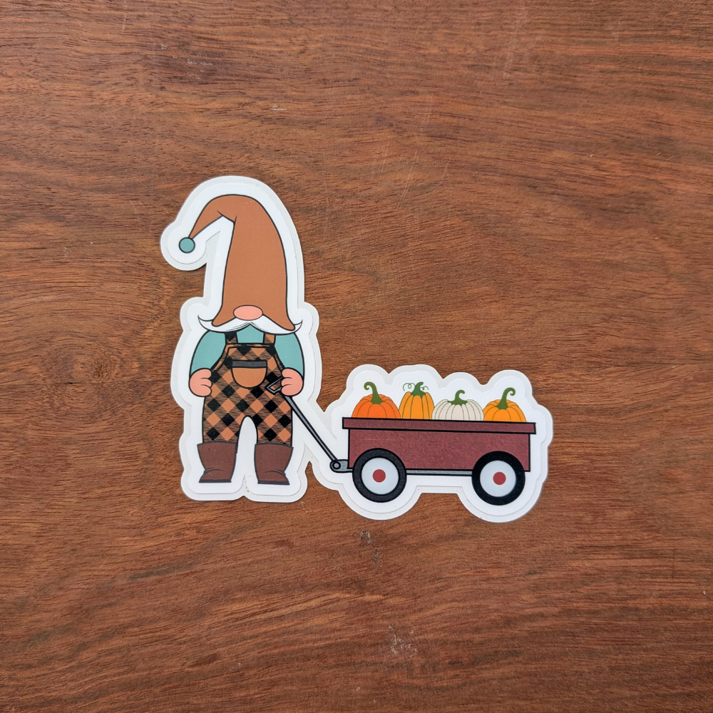A gnome sticker wearing black-and-orange buffalo plaid overalls. He is pulling a red wagon full of pumpkins.