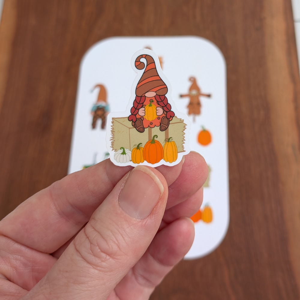 A hand holding a fall girl gnome sticker. She is sitting on a hay bale and holding a pumpkin. A fall gnome sticker sheet is on the table.