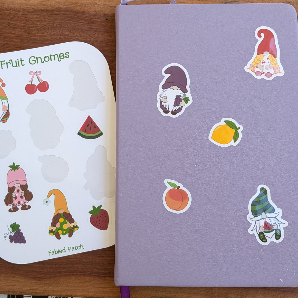 A purple journal with a cherry gnome, a grape gnome, and a watermelon gnome on the fruit. The fruit sticker sheet is seen behind the journal.