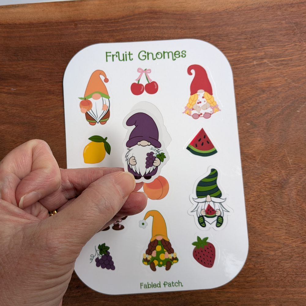 A hand holding a gnome wearing a purple hat, holding a bunch of grapes. The fruit gnome sticker sheet is behind the gnome sticker.