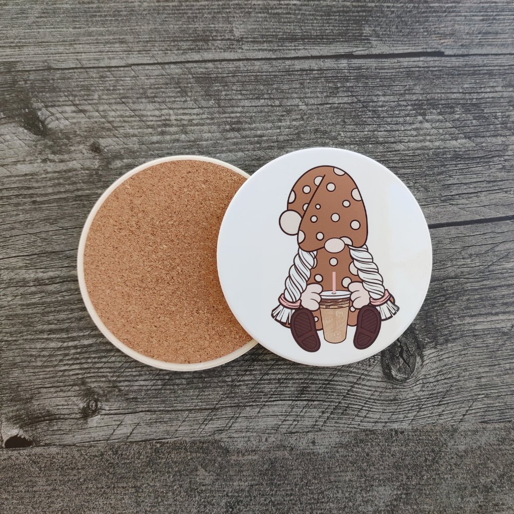 A white ceramic coaster featuring a girl gnome in a brown polka-dot dress holding an iced coffee. A back view of the coaster showing its protective cork backing.