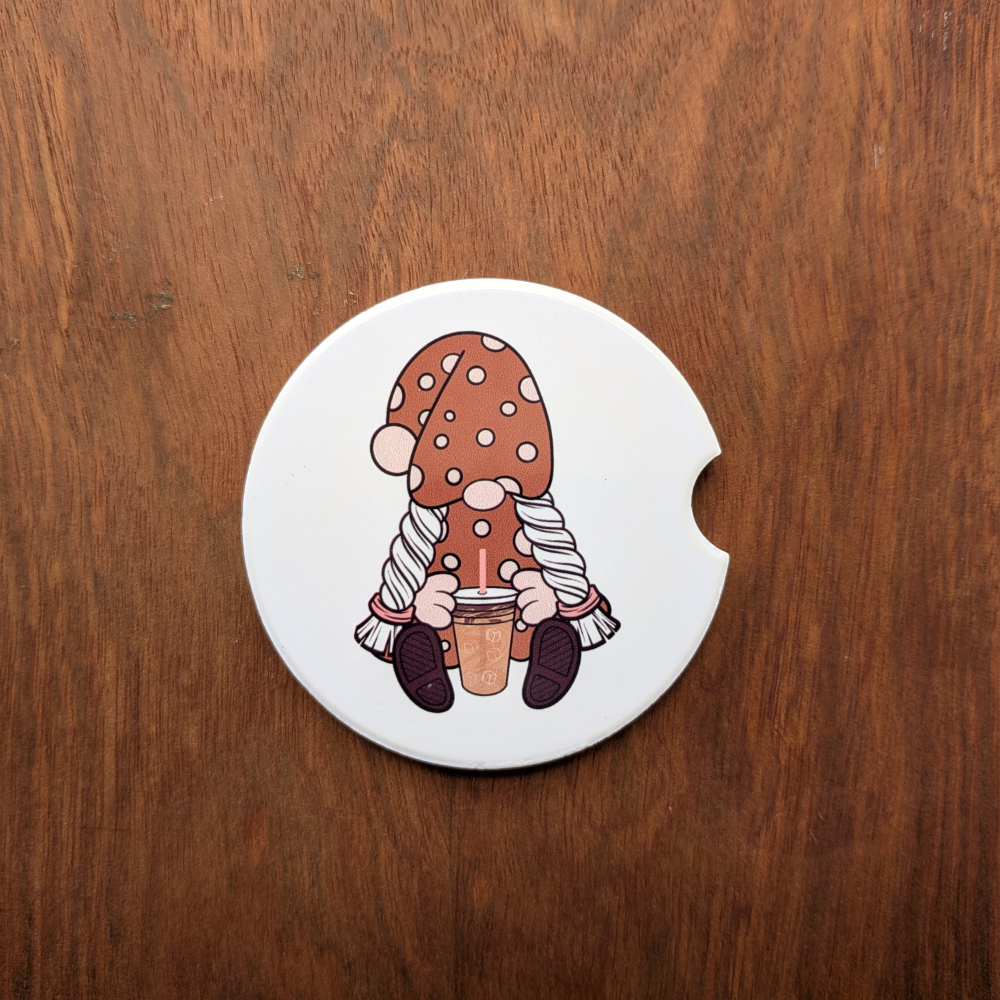 A white ceramic car coaster featuring a girl gnome holding an iced coffee. She is wearing a brown polka-dot hat and dress.
