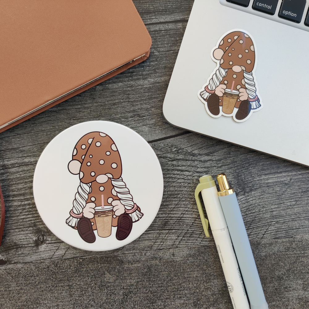 A desk featuring a laptop with a girl gnome sticker. A ceramic coaster featuring the same iced coffee gnome next to the laptop.