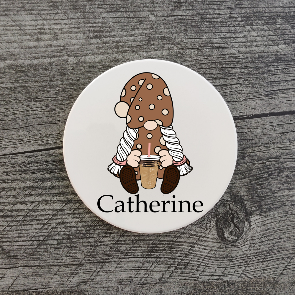 A white ceramic coaster featuring a girl gnome holding an iced coffee. The coaster is personalized with the name "Catherine."