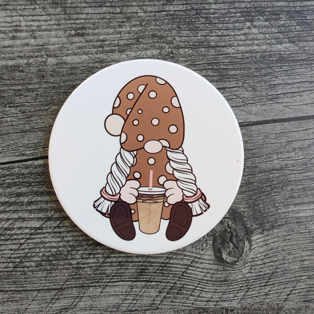 A white ceramic coaster featuring a girl gnome in a brown and cream polka-dot hat. She is holding an iced coffee.