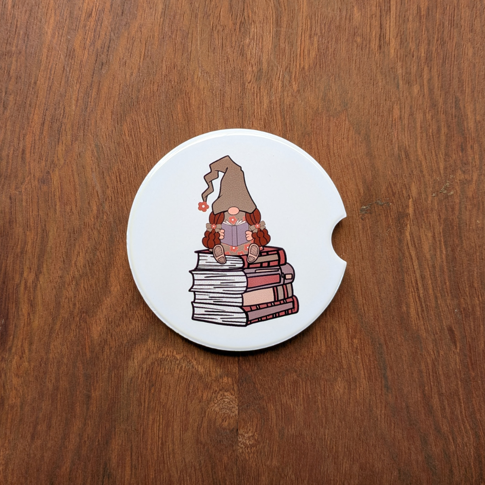 A white ceramic car coaster featuring a girl gnome in a green hat sitting on a stack of books.