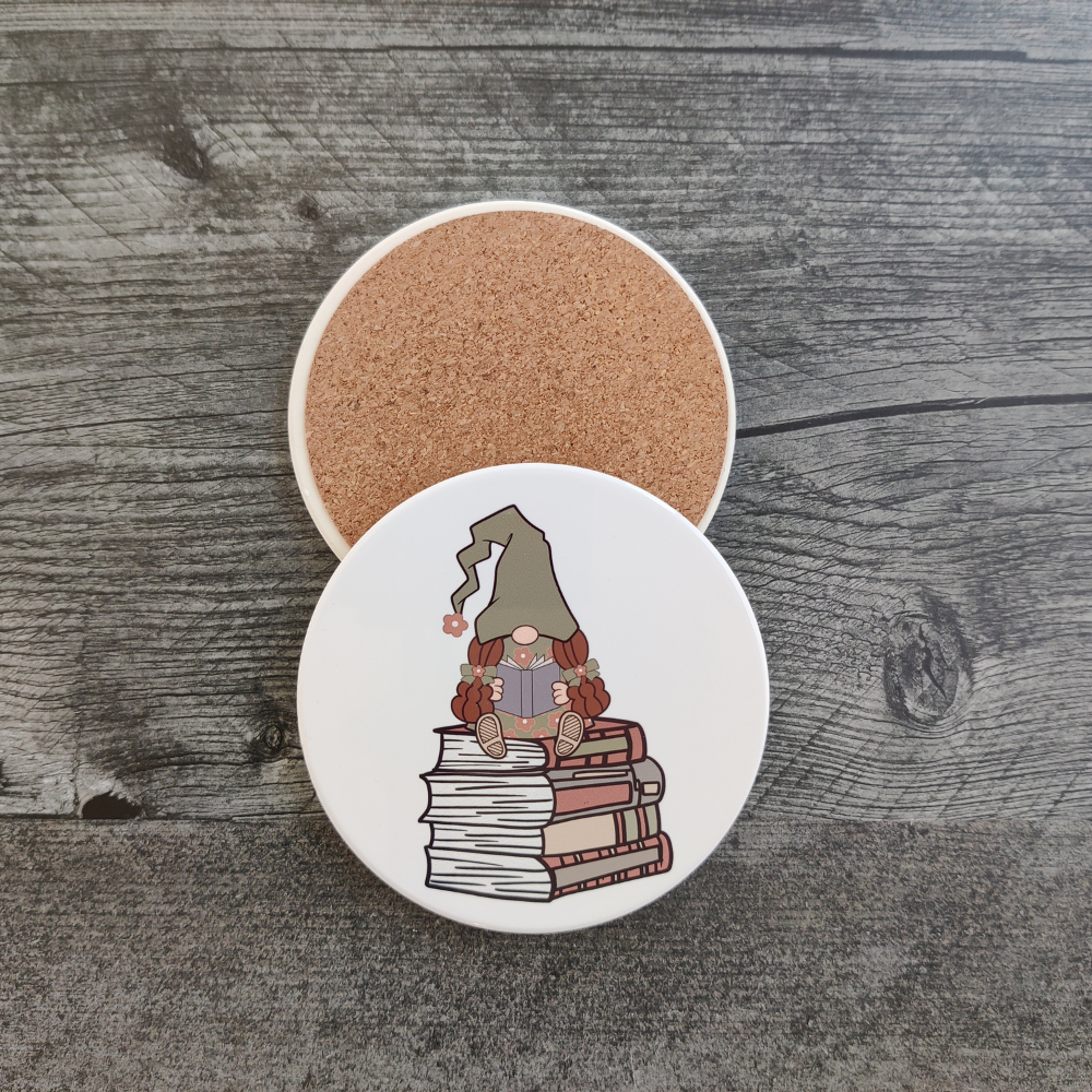 A white circular coaster featuring a girl gnome in a green hat sitting on a stack of books. A back view of the coaster showing its protective cork backing.