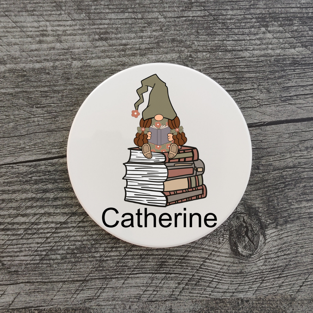 A white coaster with a girl gnome in a green hat reading a book. A girl gnome sitting on a stack of books with the name “Catherine” displayed underneath.