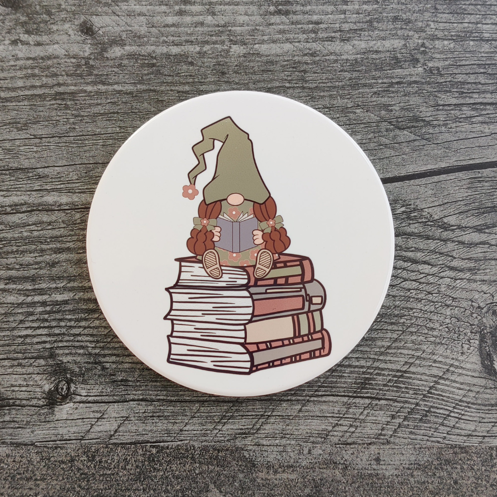 A white circular coaster featuring a girl gnome in a green hat reading a book. She is sitting on a stack of books.