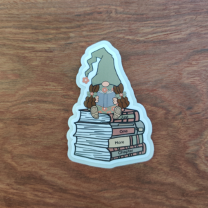 A die-cut sticker featuring a girl gnome in a green hat reading a book. She is sitting on a stack of books that reads "Just One More Chapter."