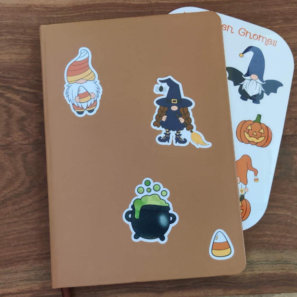 A brown journal with 3 Halloween stickers featuring a witch gnome, a candy corn, and a black cauldron. The Halloween Gnome sticker sheet is behind the journal.