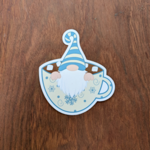 A gnome wearing a blue and cream hat sitting in a cup of hot chocolate sticker. The cup is cream with blue snowflakes.
