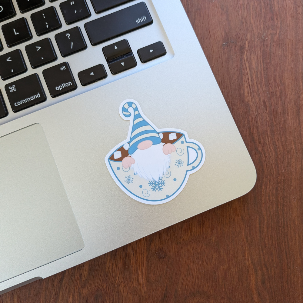 A silver laptop with a gnome sticker in the bottom-left corner. The gnome is wearing a blue and cream striped hat and sitting in a cup of hot chocolate.