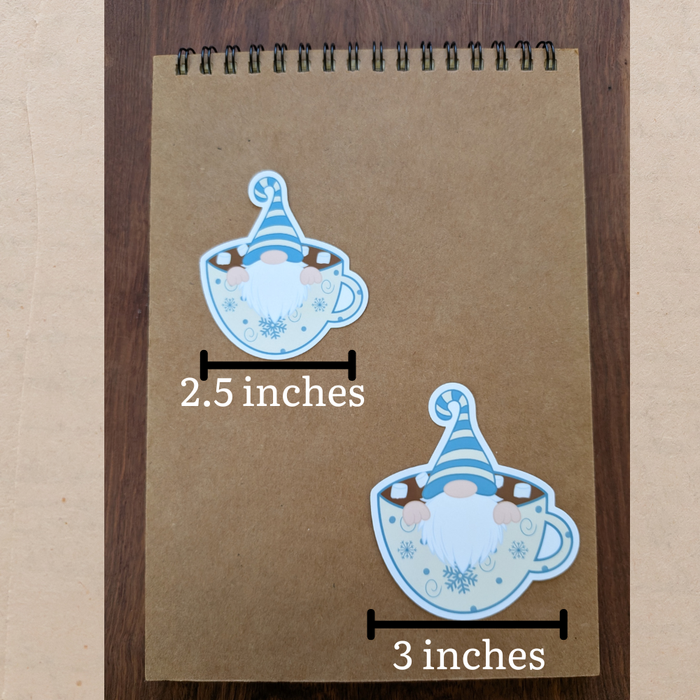 A brown spiral notebook featuring two gnomes sitting in hot chocolate stickers. The top sticker measures 2.5 inches wide. The bottom sticker measures 3 inches wide.