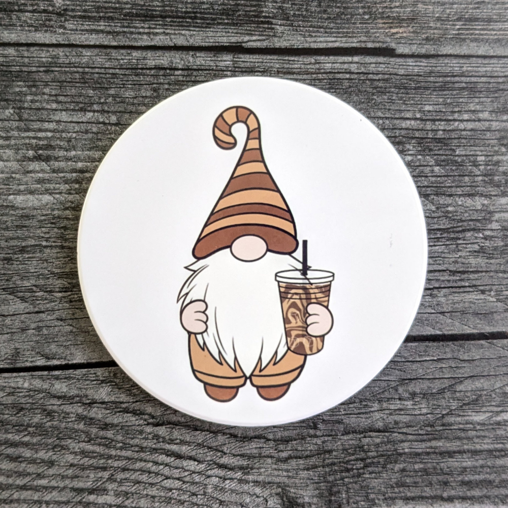 A white circular coaster featuring a gnome in a brown striped hat. He is holding an iced coffee with a black straw.