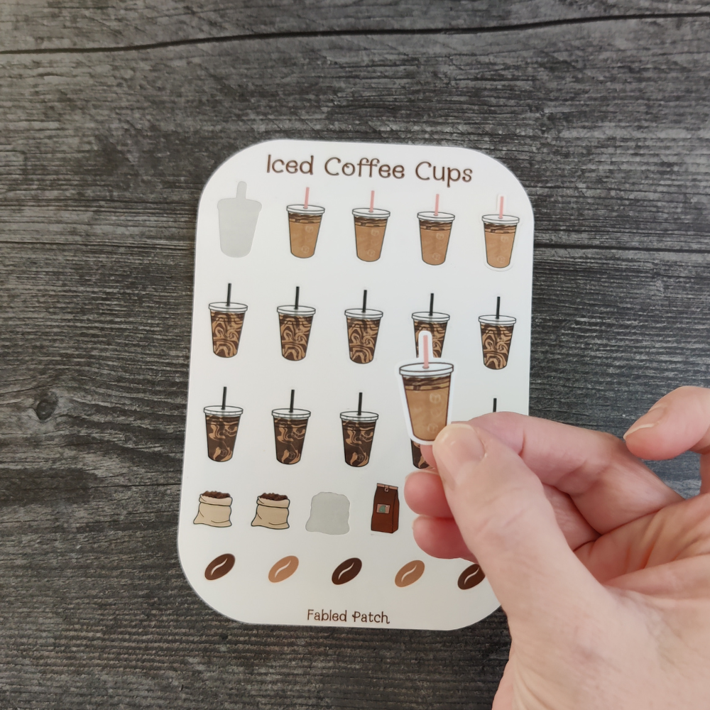 A hand holding an iced coffee sticker, with a sheet of iced coffee and coffee bean stickers in the background.