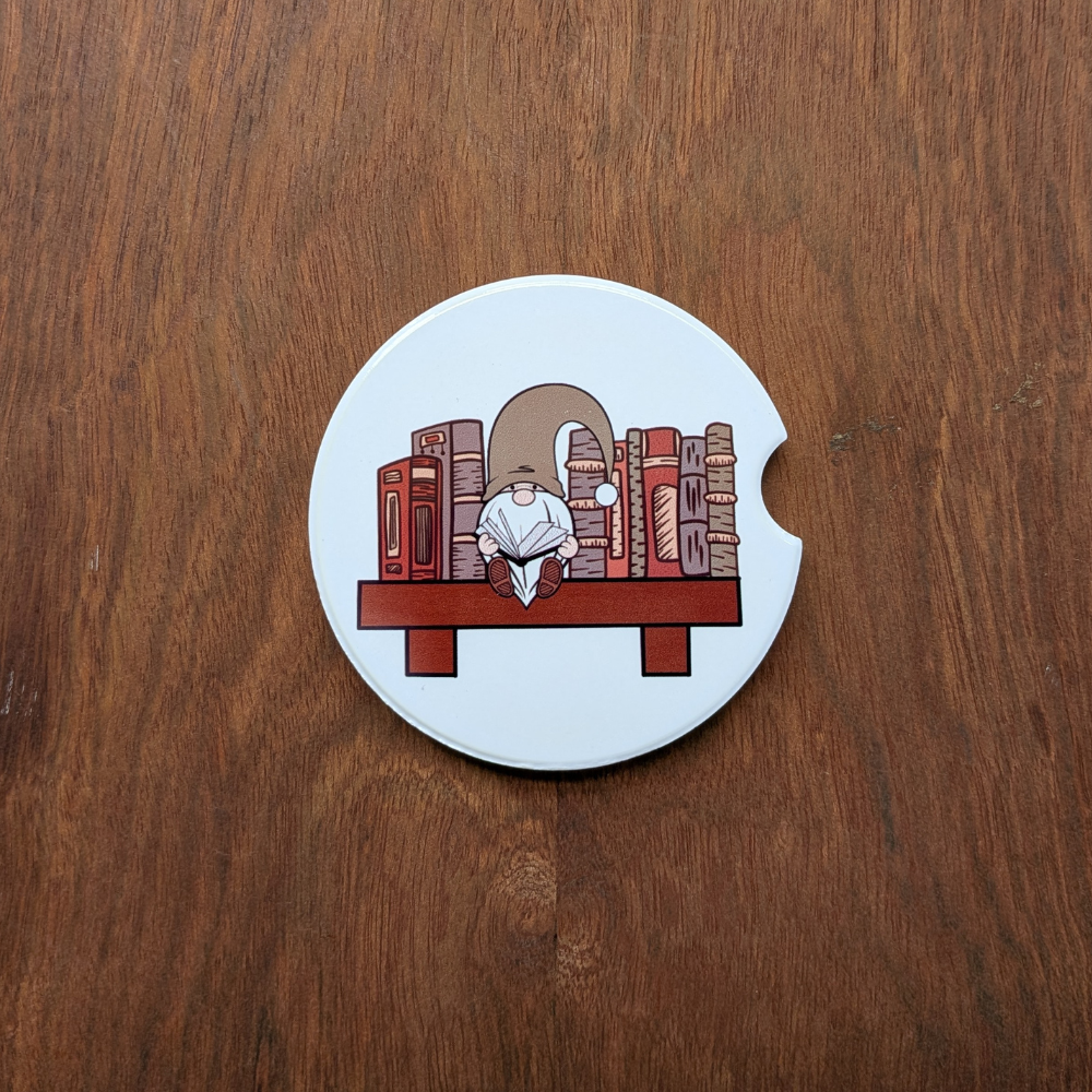 A white ceramic car coaster featuring a gnome in a green hat, sitting on a bookshelf and reading.