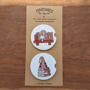 2 reading gnome car white ceramic coasters on a backing card that reads "Gnomes on the Go." One gnome is sitting on a bookshelf, and the girl gnome is sitting on a stack of books.