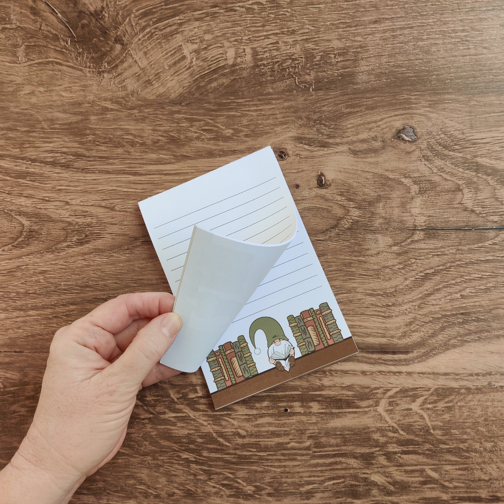 A hand flips the pages of the lined reading gnome notepad.