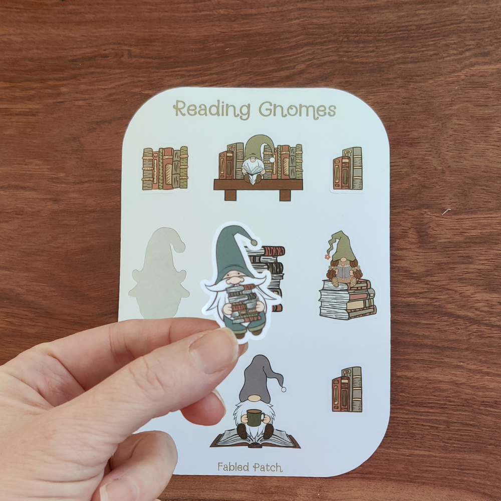 A hand holding a gnome in a blue hat that is holding a stack of books. The reading gnome sticker sheet is in the background.