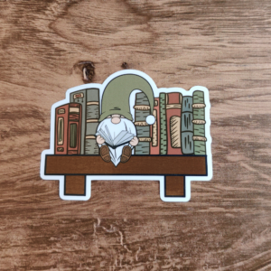 A die-cut sticker featuring a gnome in a green hat reading a book while sitting on a brown bookshelf with eight books in shades of brown, green, and red.