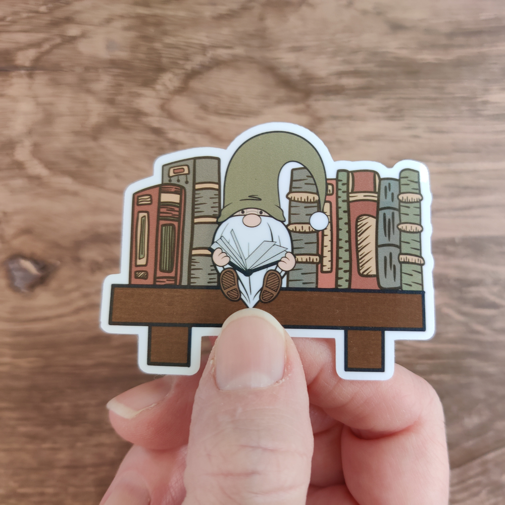 A hand holding a die-cut sticker featuring a reading gnome sitting on a bookshelf. sticker. The sticker has a clear laminate.