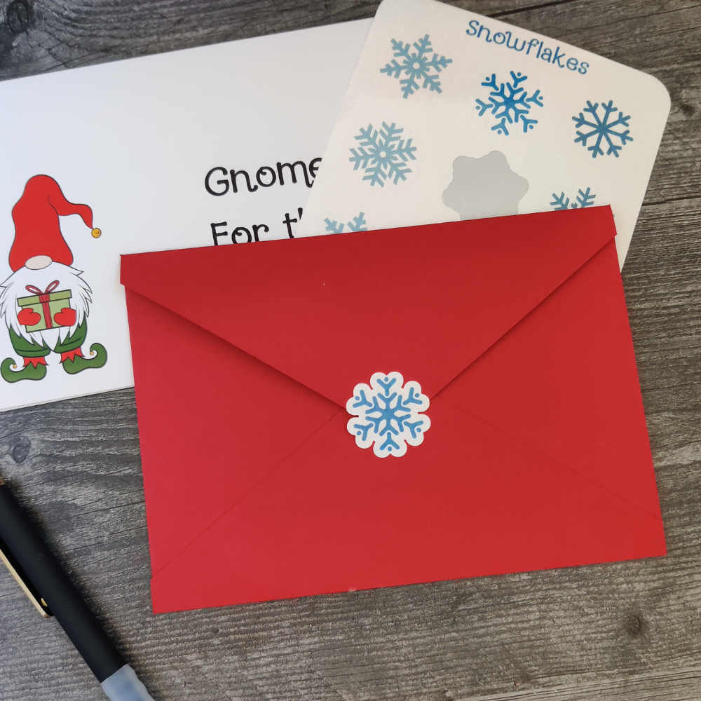 A gnome Christmas card with a red envelope on top. The envelope is closed with a blue snowflake sticker.