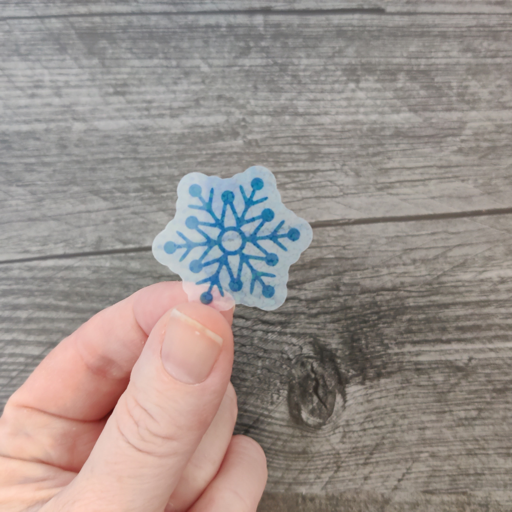 A hand holding a blue snowflake sticker with a clear background.
