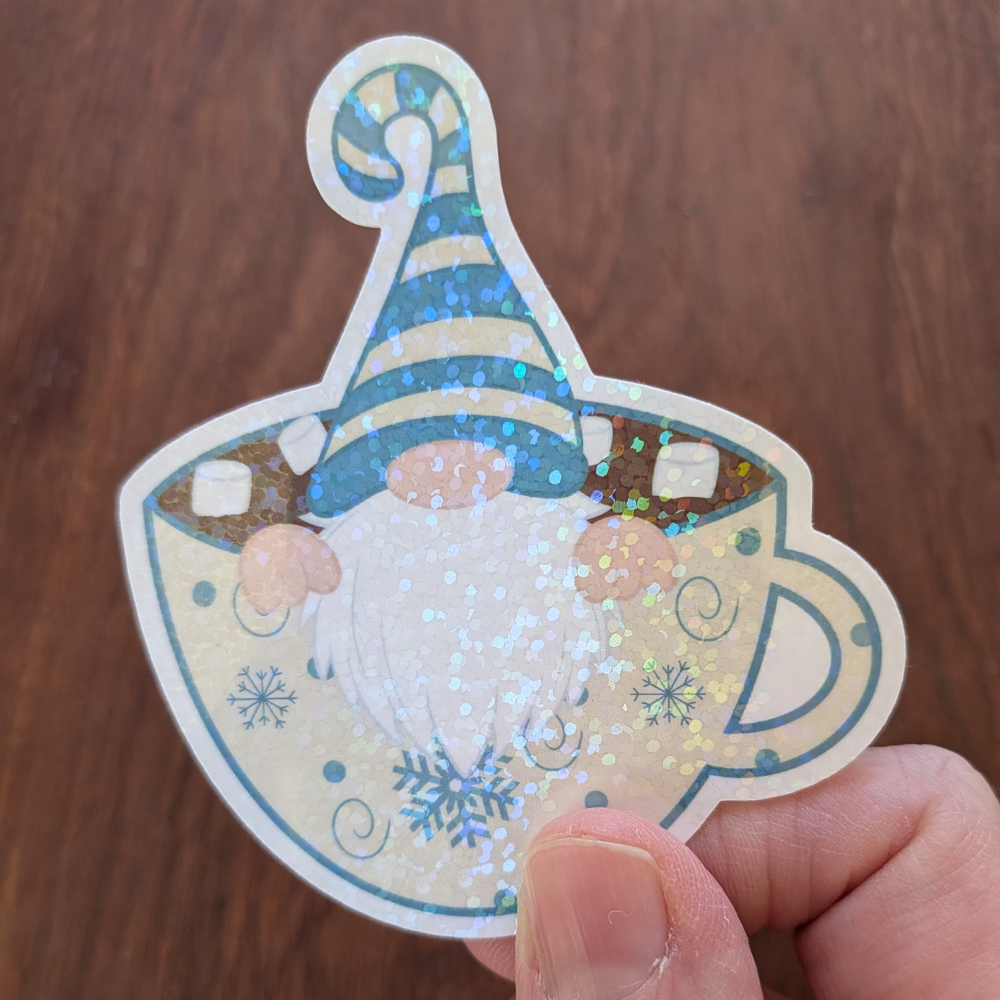 A hand holding a gnome sticker wearing a blue striped hat sitting in a cup of hot chocolate. The sticker has a sparkle laimate.