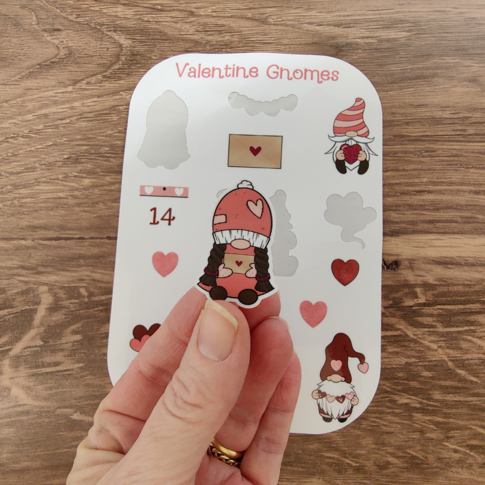 A hand holding a girl gnome sticker wearing a pink hat decorated with hearts, with a Valentine’s Day gnome sticker sheet displayed in the background.